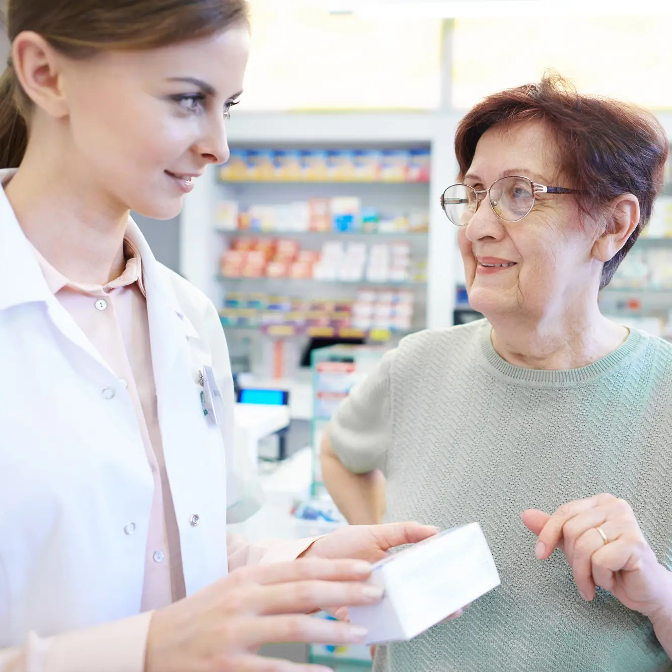 Eine Apothekenangestellte mit Medikament in der Hand und eine ältere Kundin mit Brille